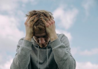 Young man in gray hoodie holds head in frustration, set against cloudy sky.